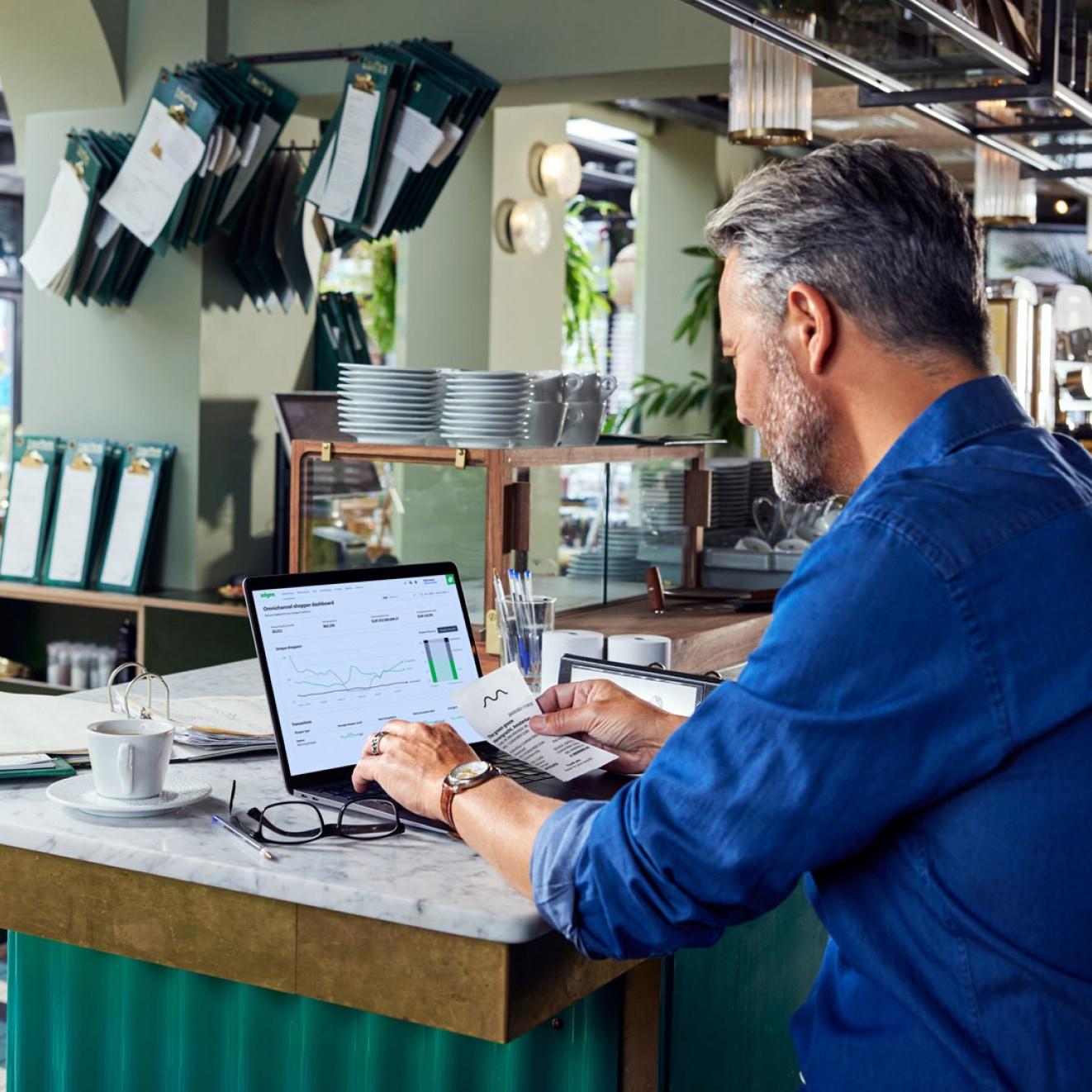 Mann in Café analysiert Verkaufsdaten auf Laptop-Bildschirm mit sichtbarer Adyen-Analyse.