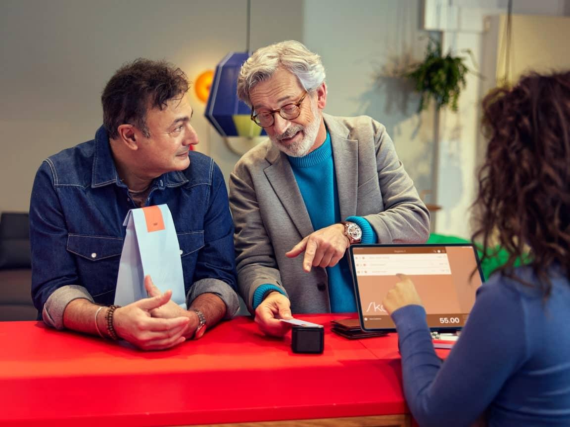 Deux hommes discutant une transaction avec une caissière utilisant un terminal de paiement numérique sur une tablette.